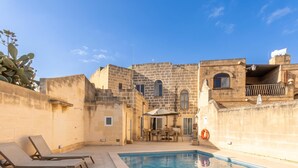 Un área junto a la piscina con sillones de descanso, una mesa de comedor y un edificio de piedra con ventanales de arco.