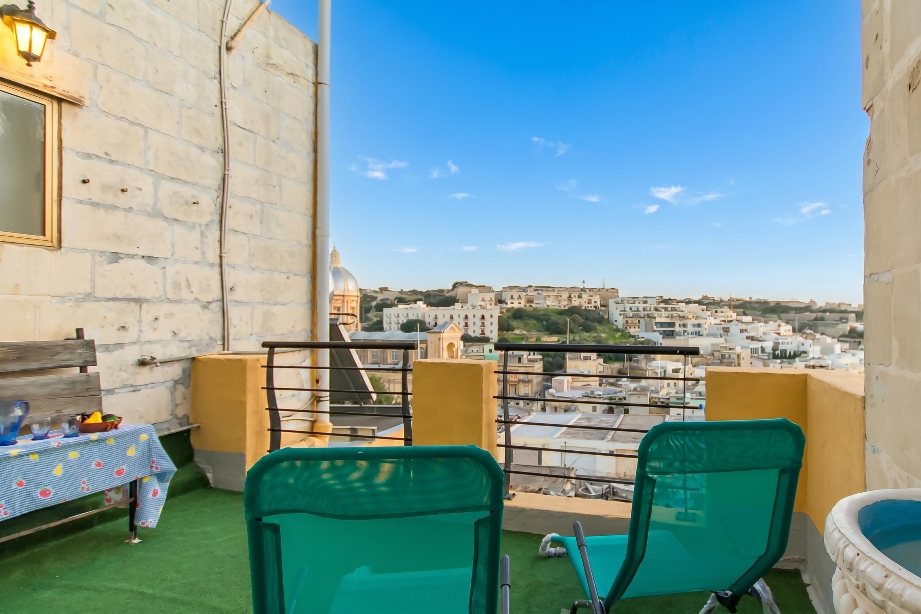 A balcony with a view of a cityscape, a table with a tablecloth, and two chairs.
