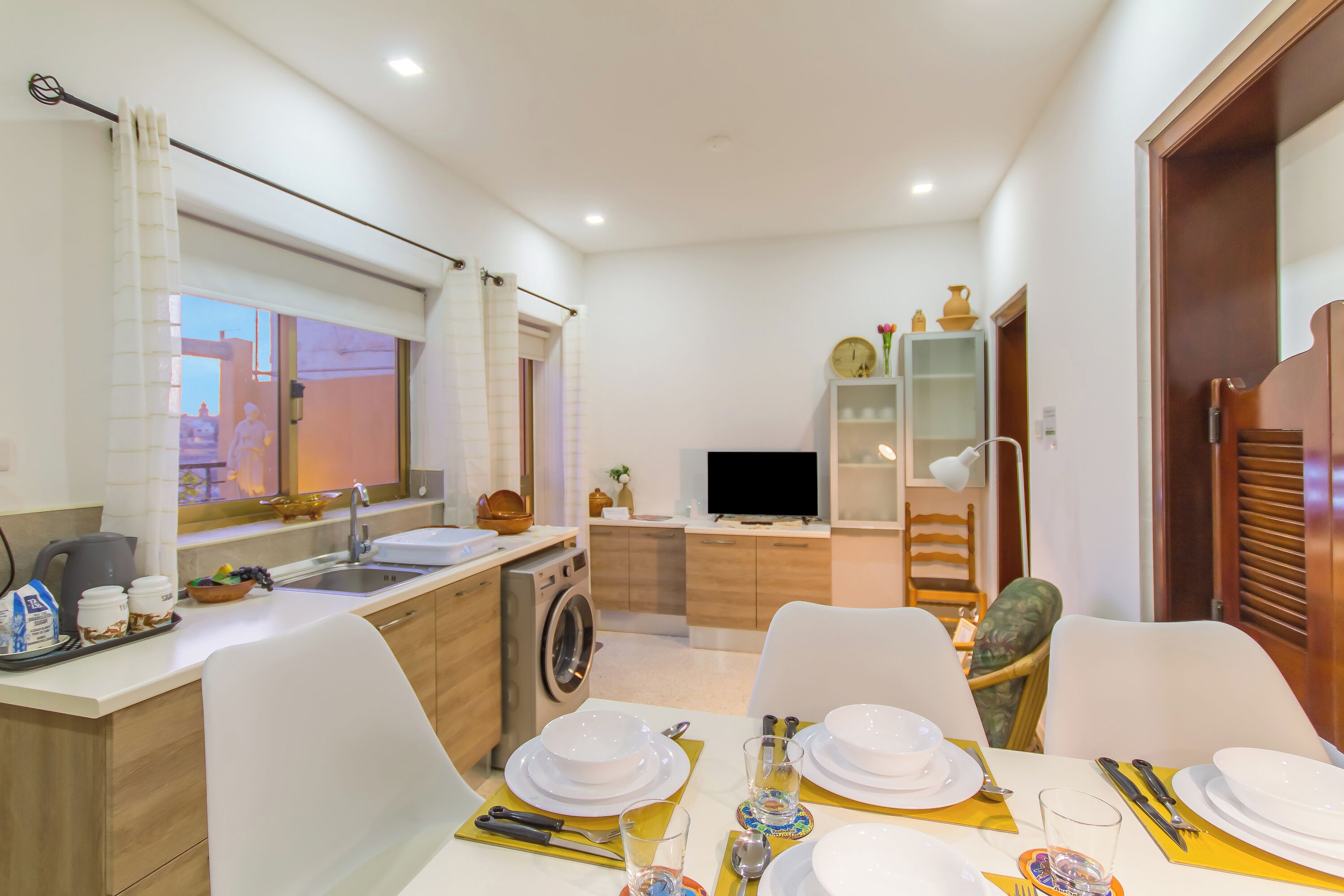 A compact kitchen with a washing machine, a dining area with a table set for four, and a TV in the background.