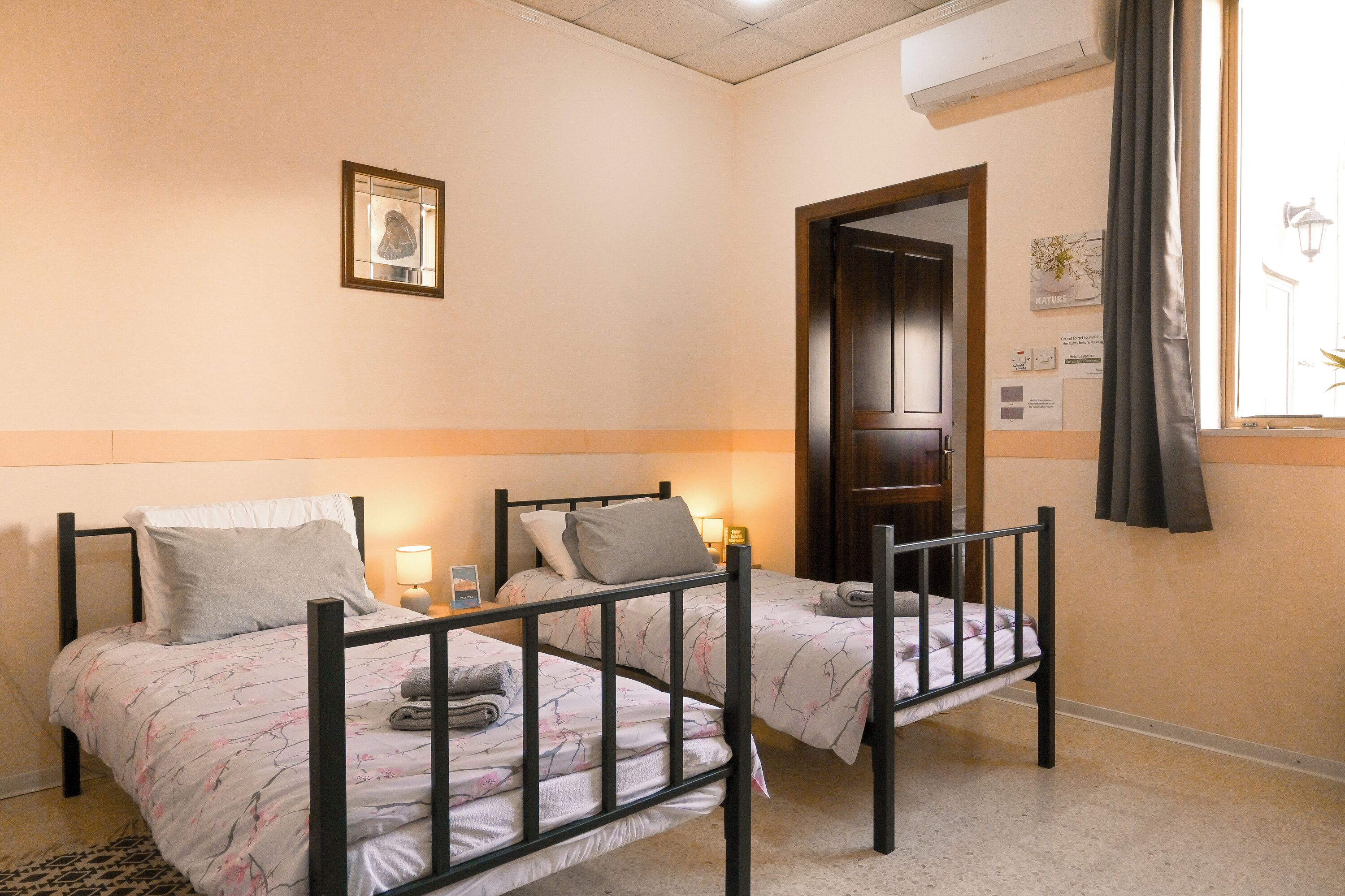 Una habitación con dos camas individuales, una mesita con lámpara, un cuadro en la pared y una puerta que da a otra estancia.