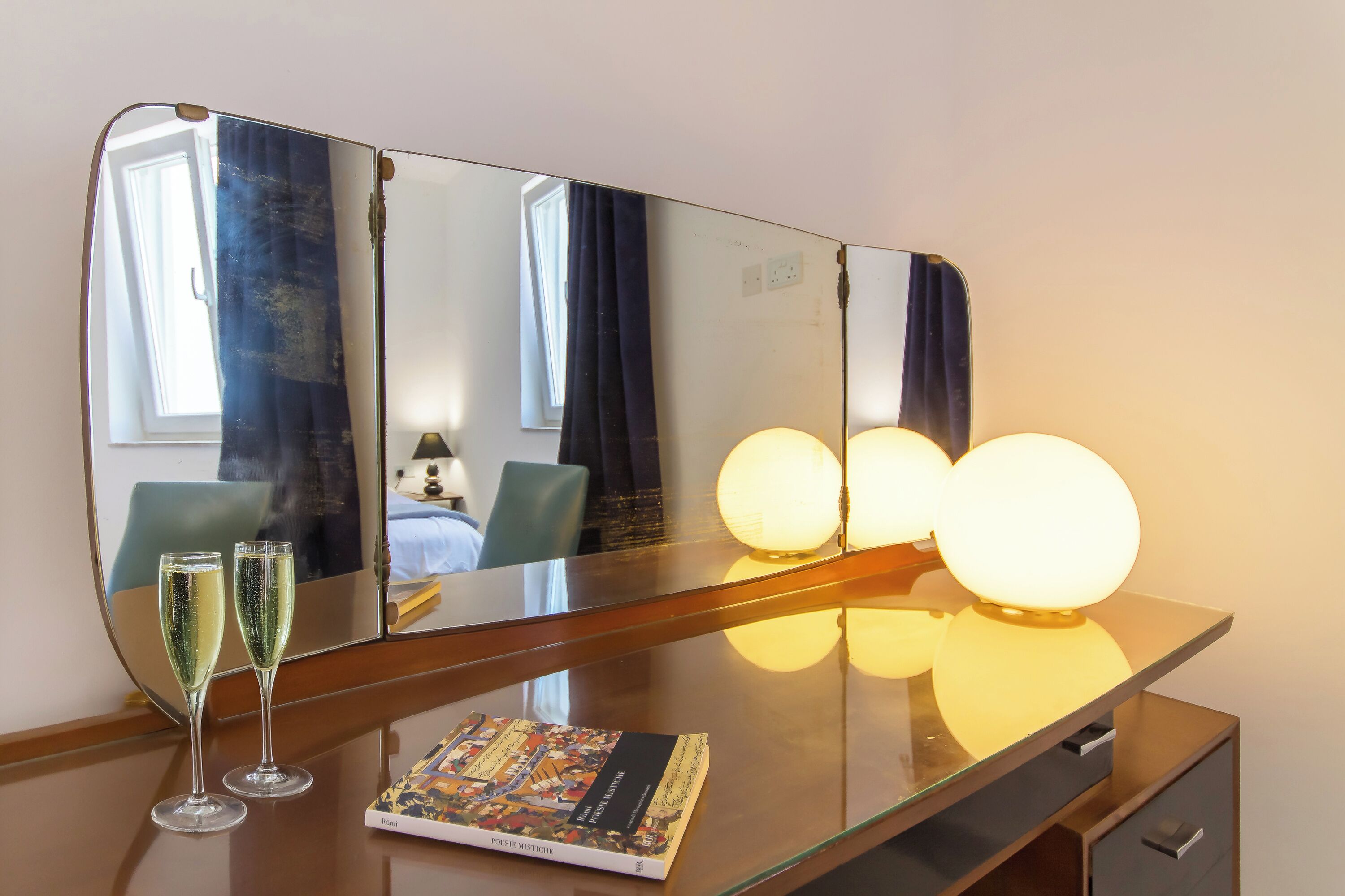 Une chambre d’hôtel avec un grand miroir, deux coupes à champagne, un livre et trois lampes murales sphériques.