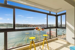 Quarto com grandes janelas com vista para um areal marítimo e paisagem urbana. Há dois taburetes amarelos e uma pequena mesa com frutas e um bule.