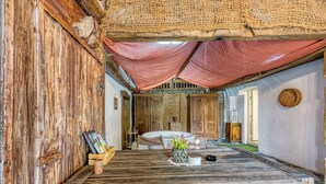 A rustic room with wooden walls and a large wooden table. There is a bed with a white mattress and a wooden headboard. A small wooden cabinet and a wicker basket are visible.