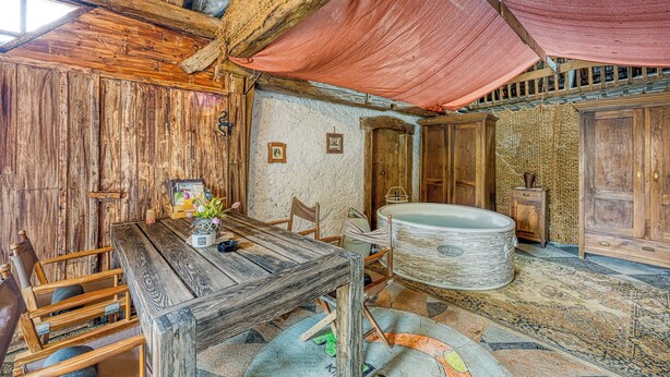 A rustic room with wooden walls, a large wooden table, chairs, a bathtub, and a patterned rug.