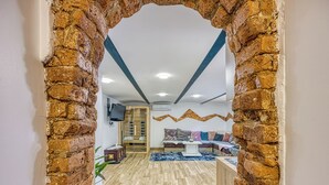 A room with a brick archway, wooden flooring, a sofa with cushions, a small table, and a TV mounted on the wall.