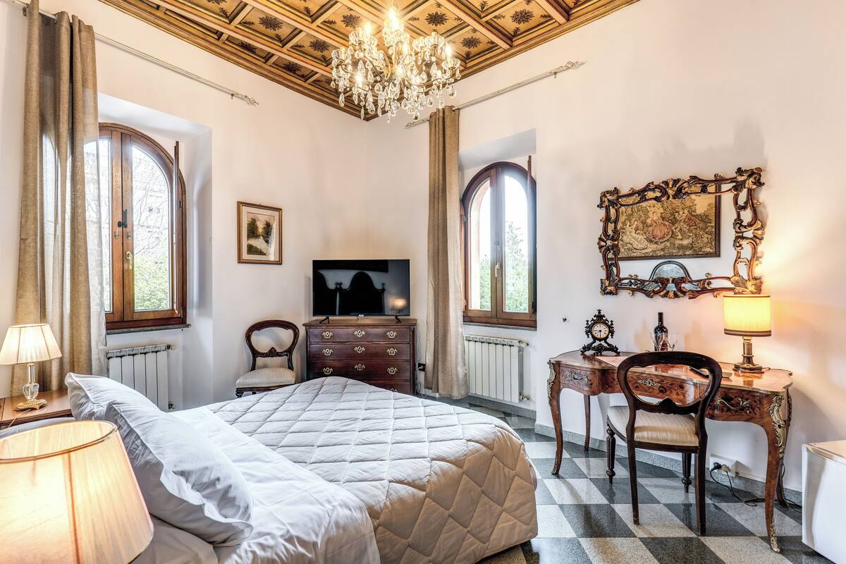 A bedroom with a bed, dresser, desk, chair, television, chandelier, and large windows.