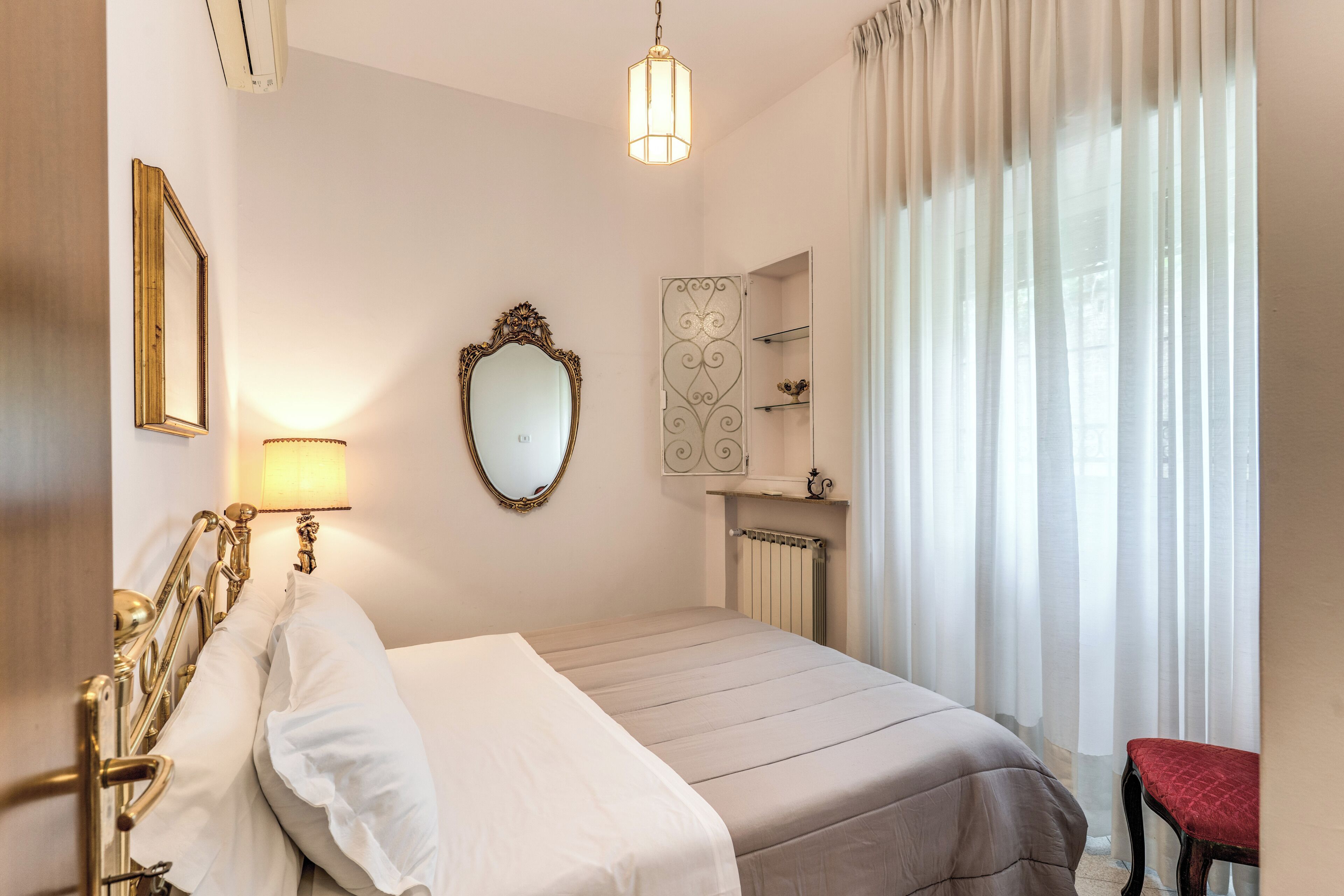 A hotel room with a bed, a mirror, a lamp, a radiator, and a window with curtains.