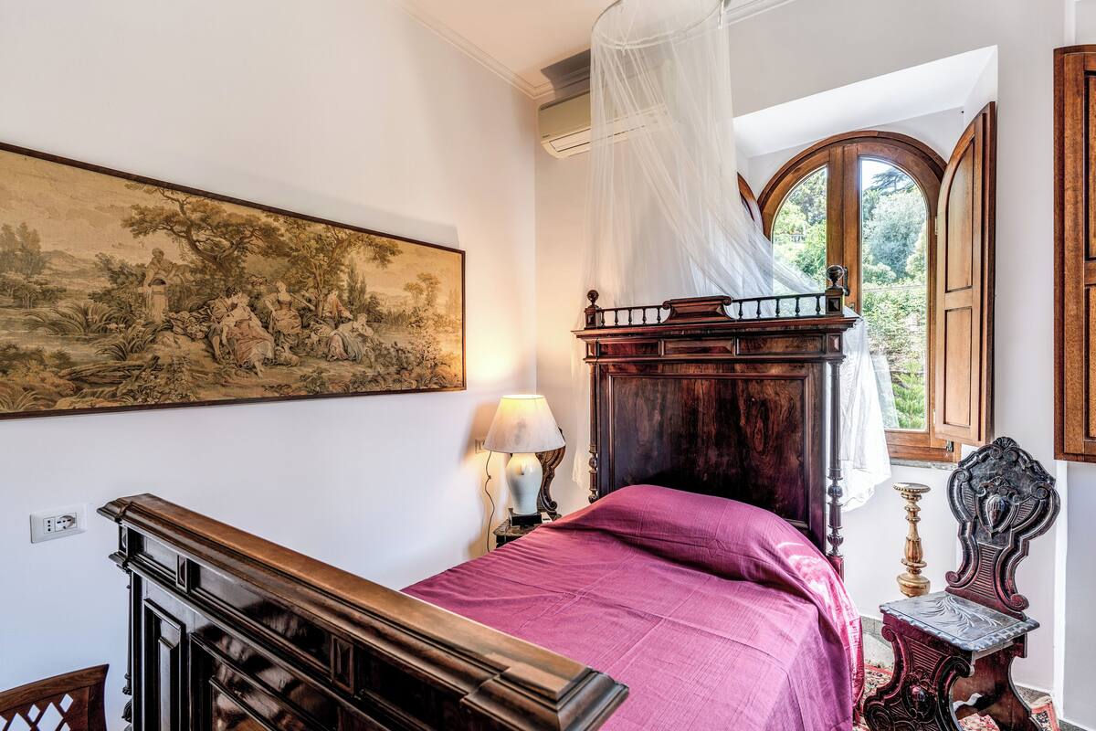 A bedroom with a bed, a nightstand, a lamp, a wooden chair, a painting, and a window with a view of greenery.