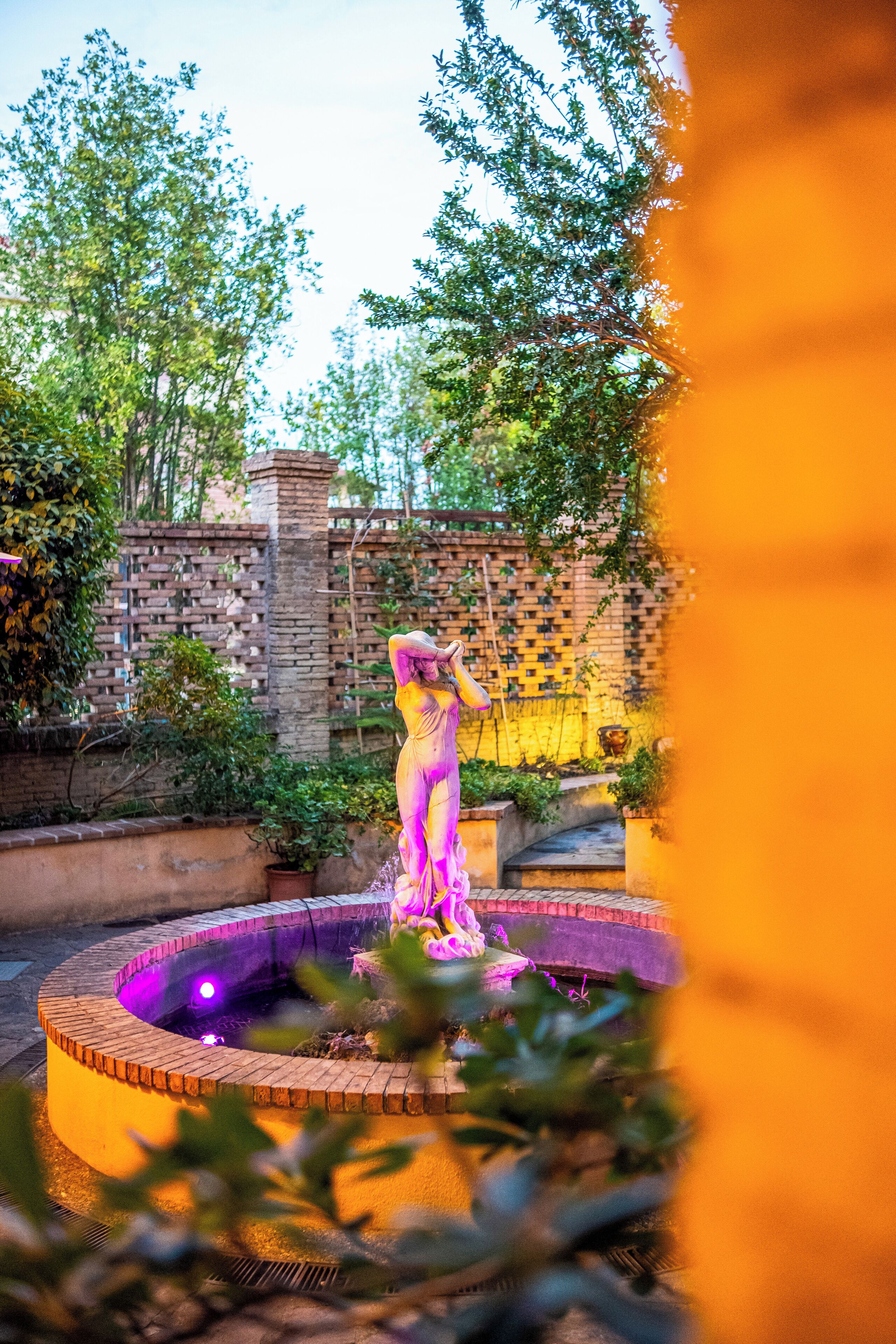 A statue in a circular water feature with a brick wall and greenery in the background.