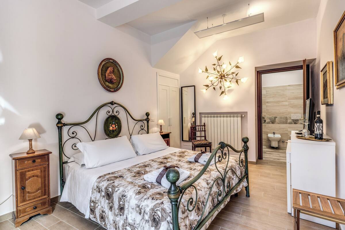 A bedroom with a bed, bedside tables, a mirror, a chandelier, and a bathroom visible through an open door.