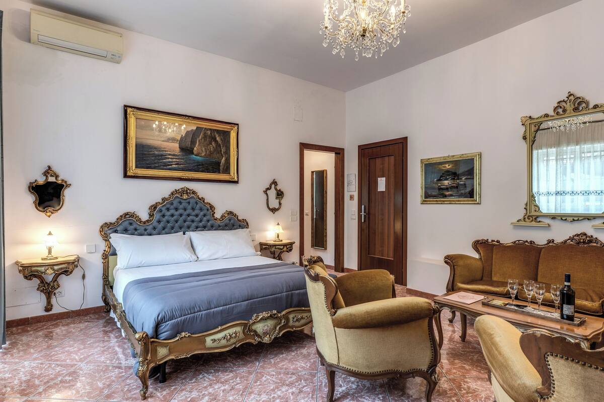 A bedroom with a large bed, a sitting area with a sofa and armchairs, a chandelier, and a painting on the wall.