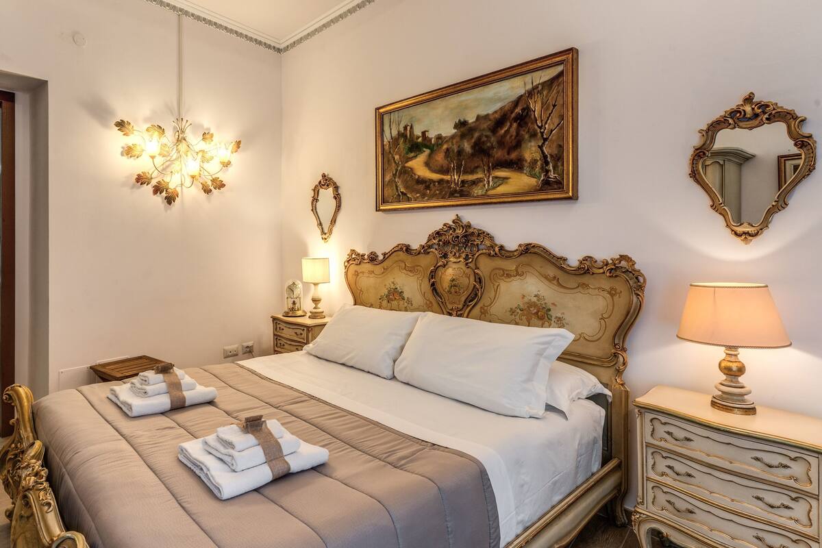 A well-appointed bedroom with a large bed, ornate headboard, a painting, a mirror, and a nightstand with a lamp.