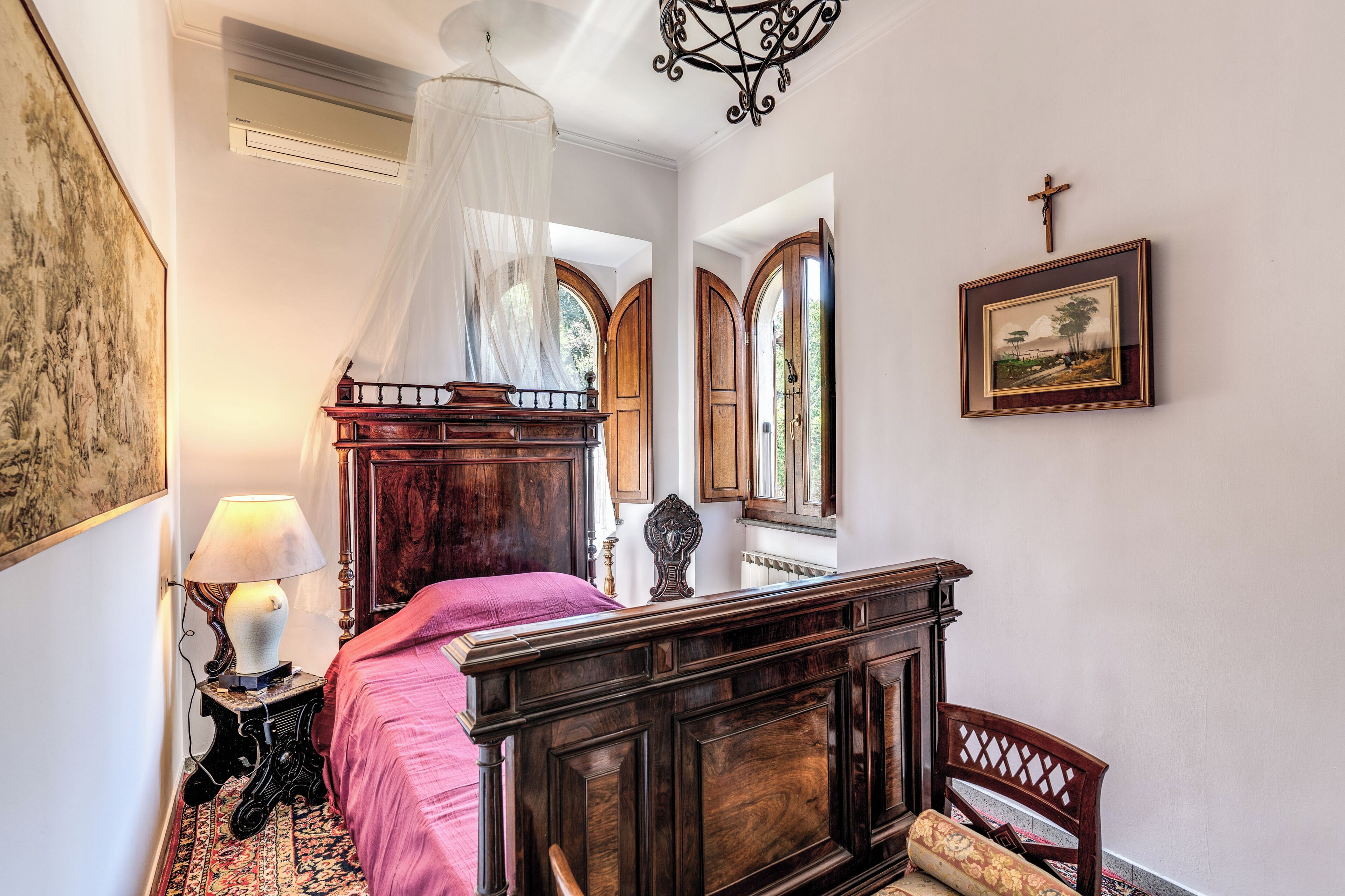 A bedroom with a wooden four-poster bed, a bedside table with a lamp, a red bedspread, and a large window with a view of greenery.