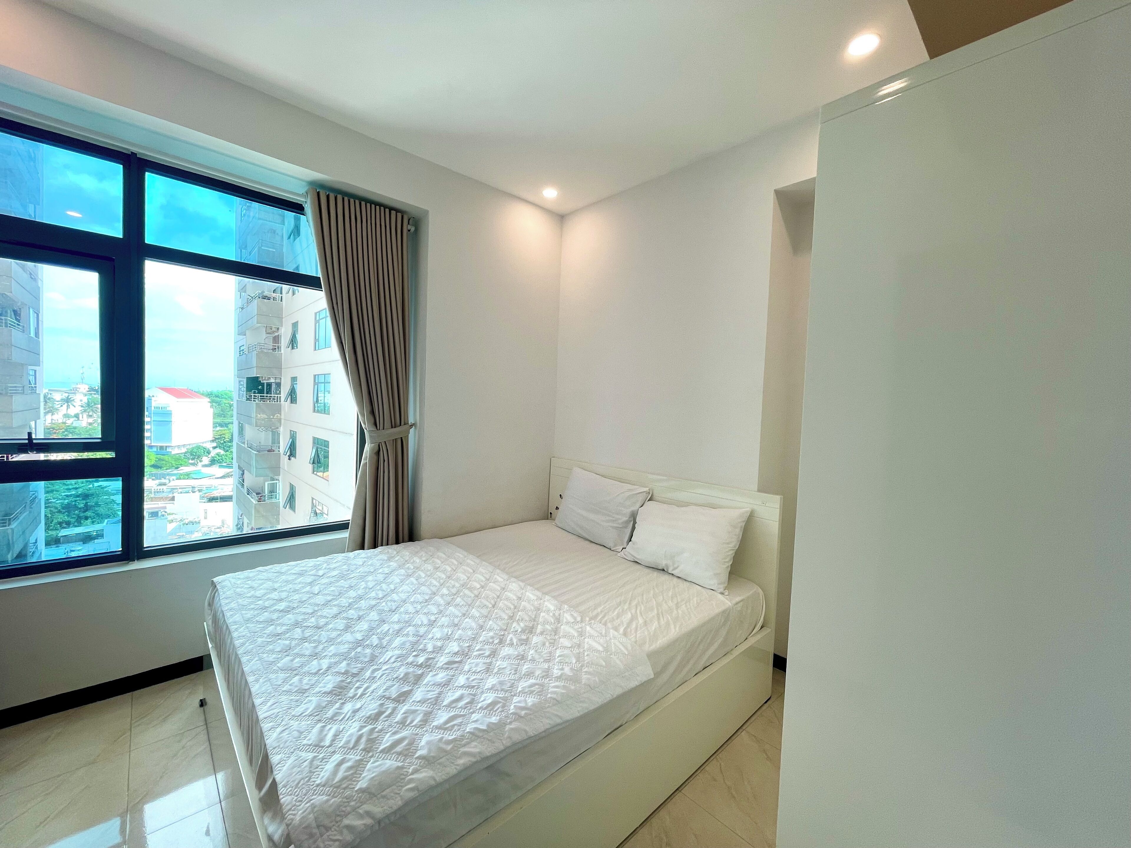 A modern hotel room with a large window, a bed with white bedding, and a view of the cityscape.