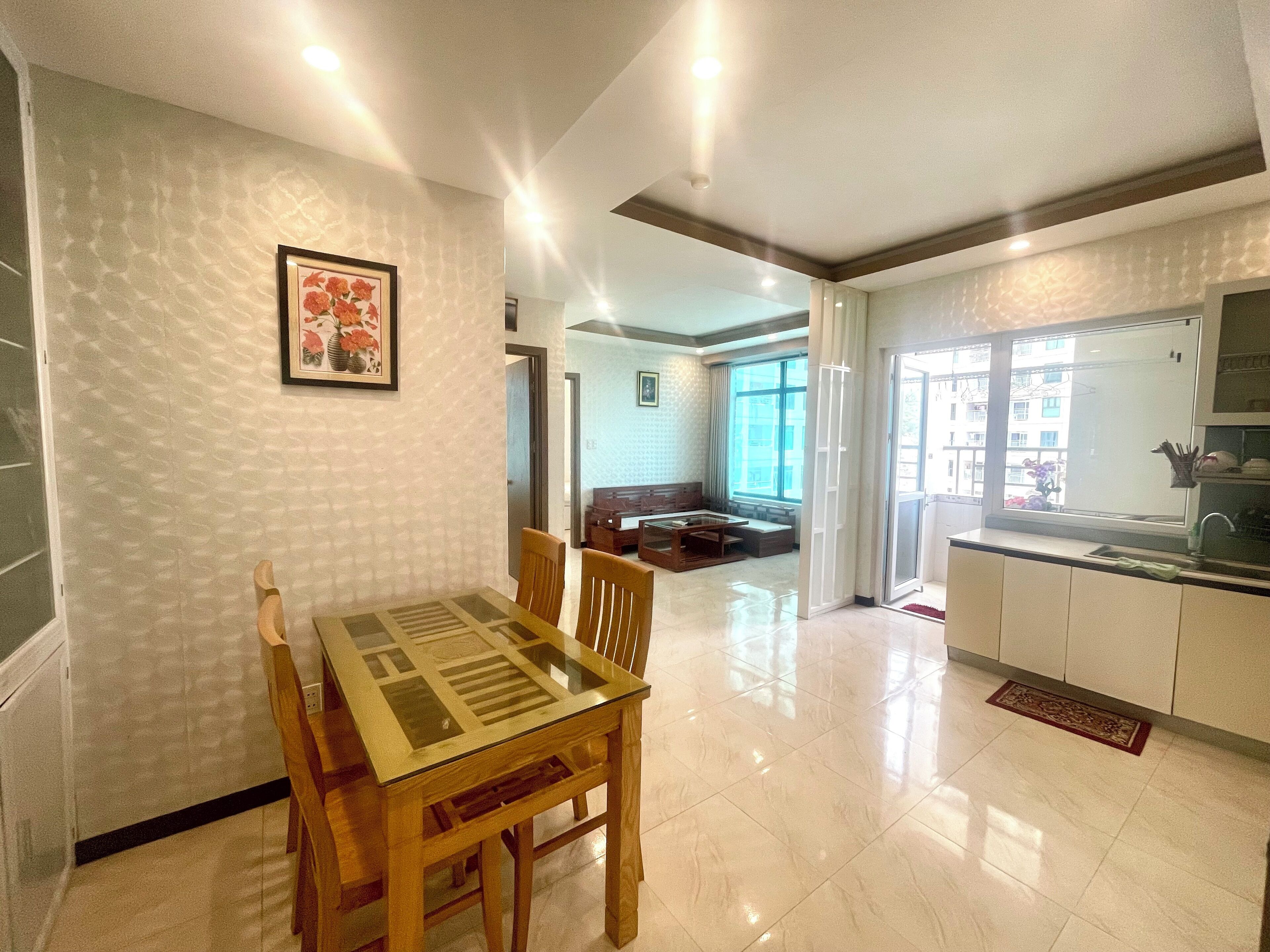 Un comedor con mesa de vidrio y sillas de madera, una cocina con lavamanos y armarios, y una sala de estar con sofá y ventana.