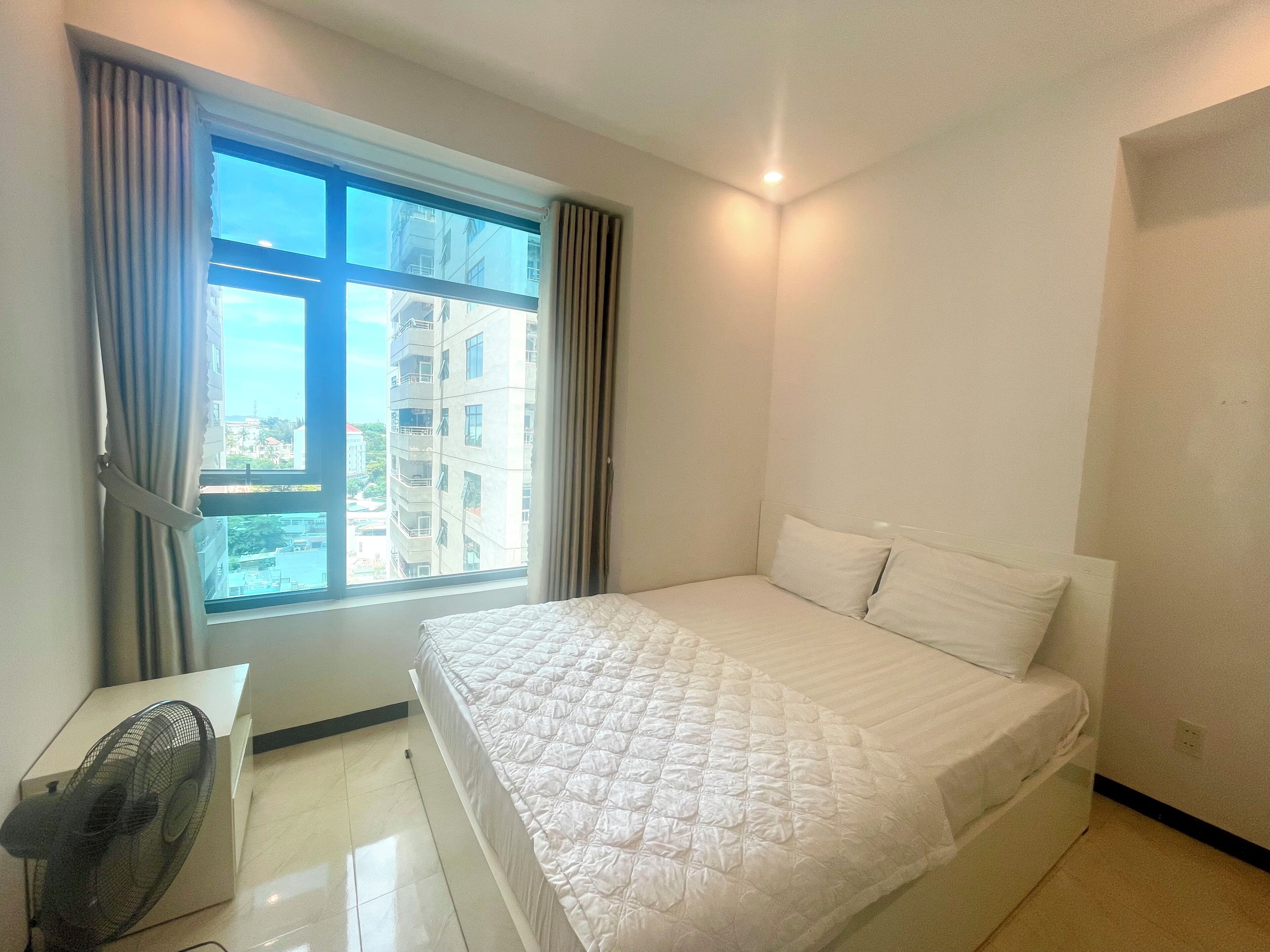 A bedroom with a bed, a window with curtains, a standing fan, and a view of buildings outside.
