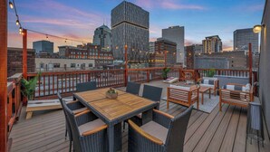 A rooftop deck with outdoor furniture, a wooden table, and a view of the city skyline at dusk.