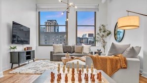 A modern living room with a chessboard, a sofa, a television, and large windows with a city view.
