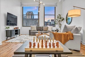 A modern living room with a chessboard, a sofa, a television, and large windows with a city view.