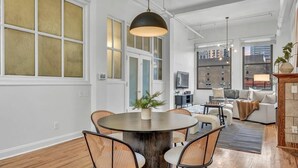 A modern living room with a round wooden table, wicker chairs, a hanging light fixture, and a view of the city through large windows.