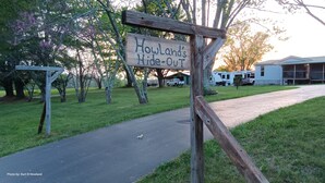 Ein hölzernes Schild mit der Aufschrift 'Howland's Hide-Out', ein gepflasterter Weg führt zu Wohnmobilen und einem Gebäude im Hintergrund.