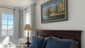 Ein Hotelzimmer mit einem großen Bett, einem Gemälde eines historischen Gebäudes, einem Fenster mit Vorhängen und einer Lampe auf einem Nachttisch.