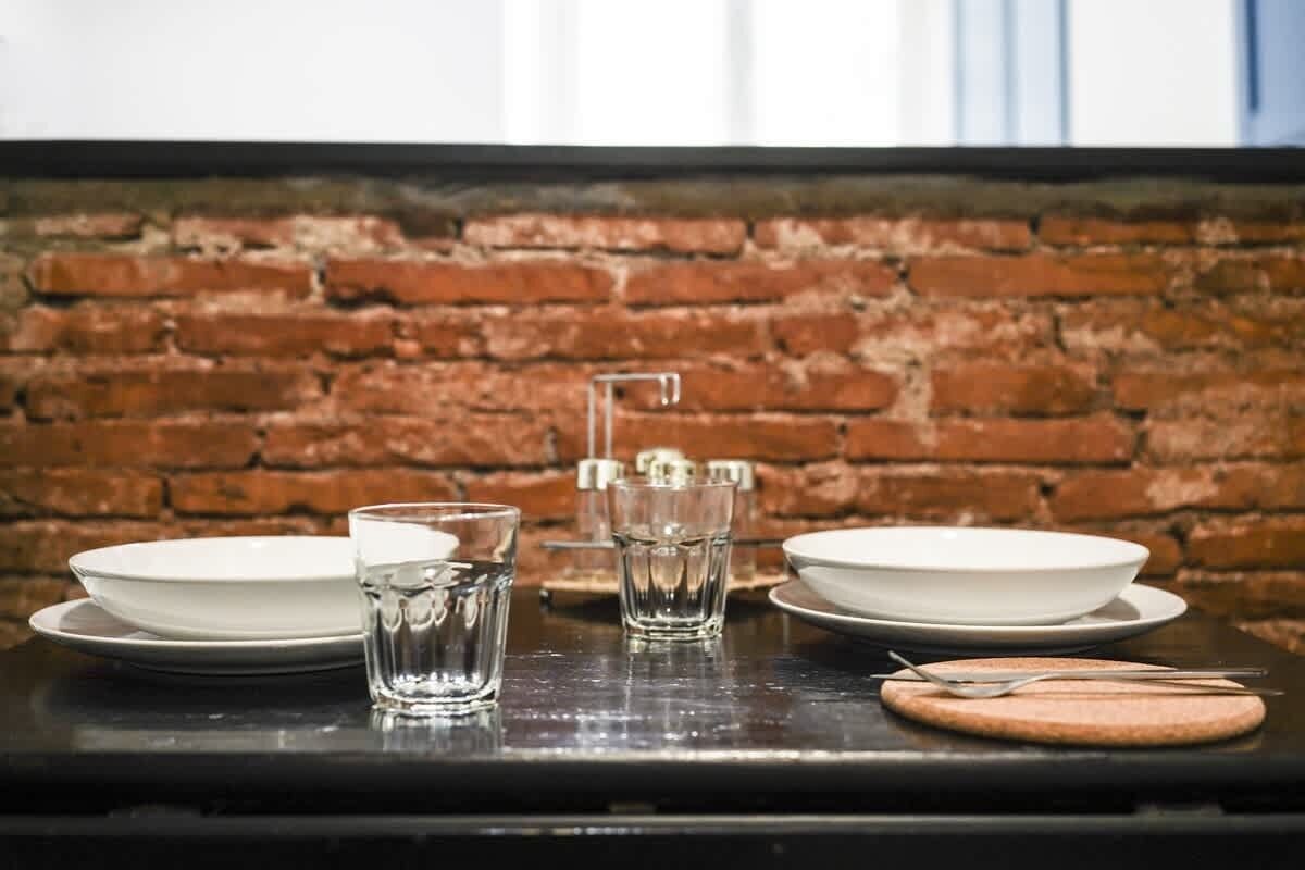 Una mesa preparada con platos, vasos y cubiertos frente a una pared de ladrillo.