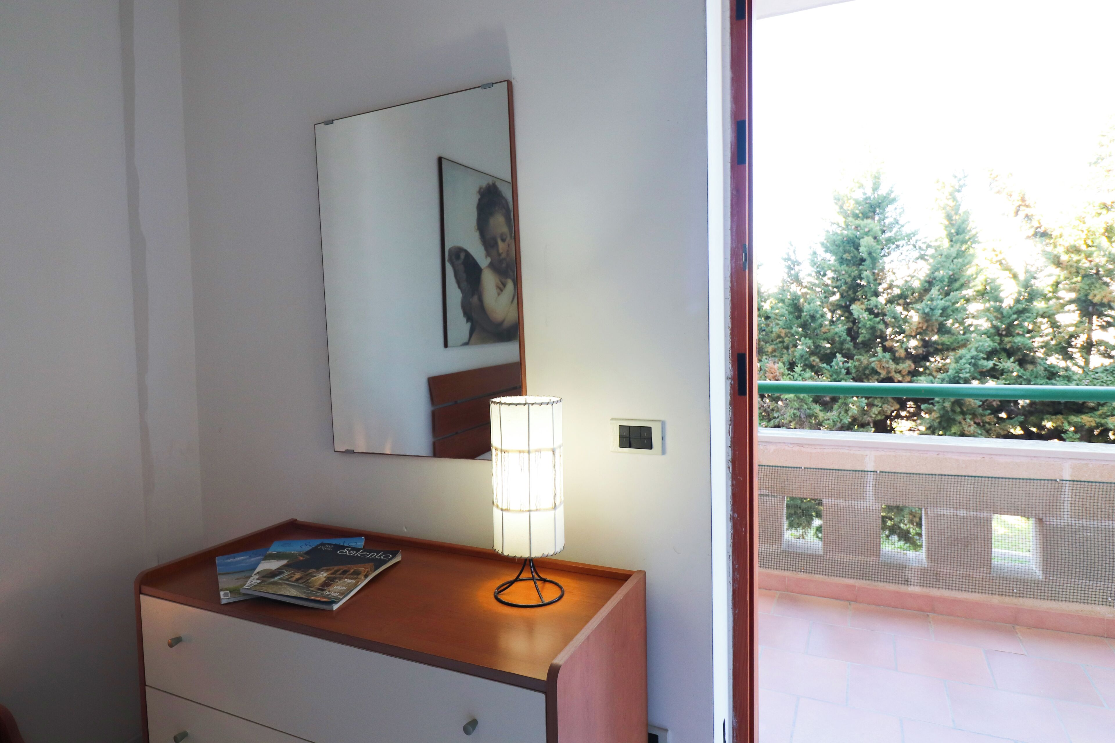 Una habitación con mesita de noche de madera, una lámpara, un espejo y una ventana con vista a árboles.
