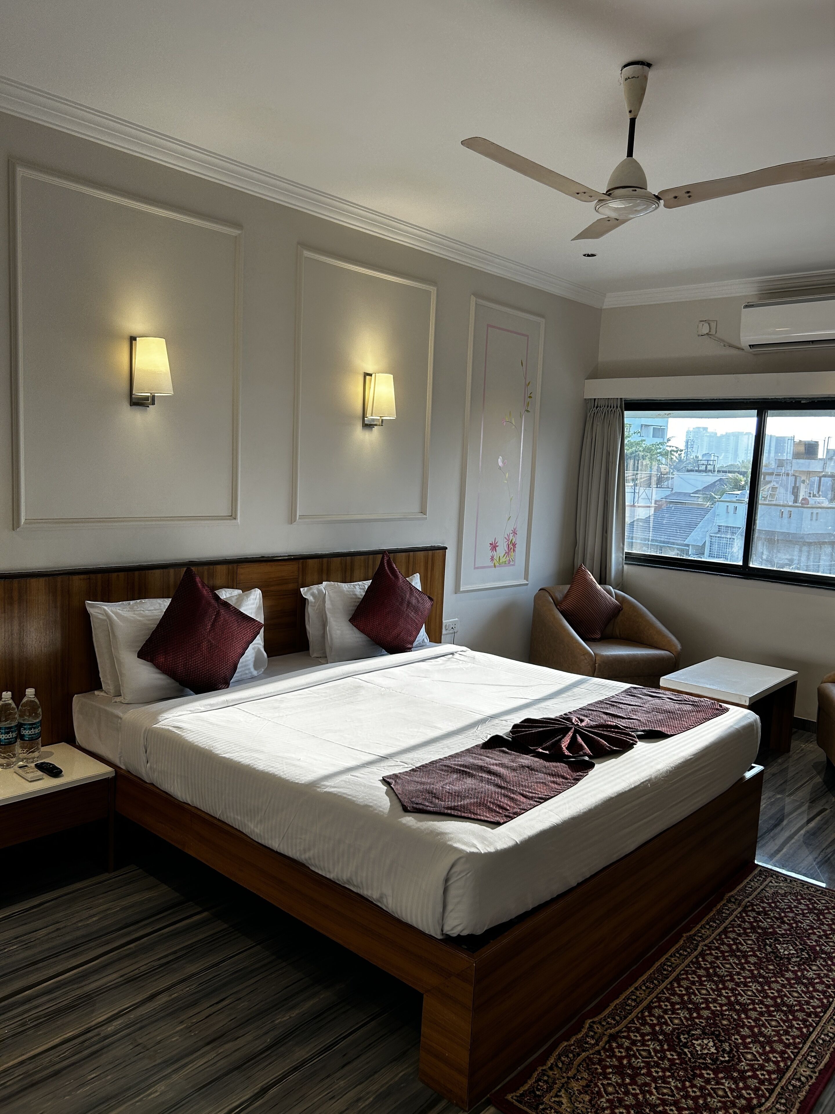 A hotel room with a large bed, two bedside tables, a ceiling fan, wall-mounted lights, a window with a view of buildings, and a wooden floor.