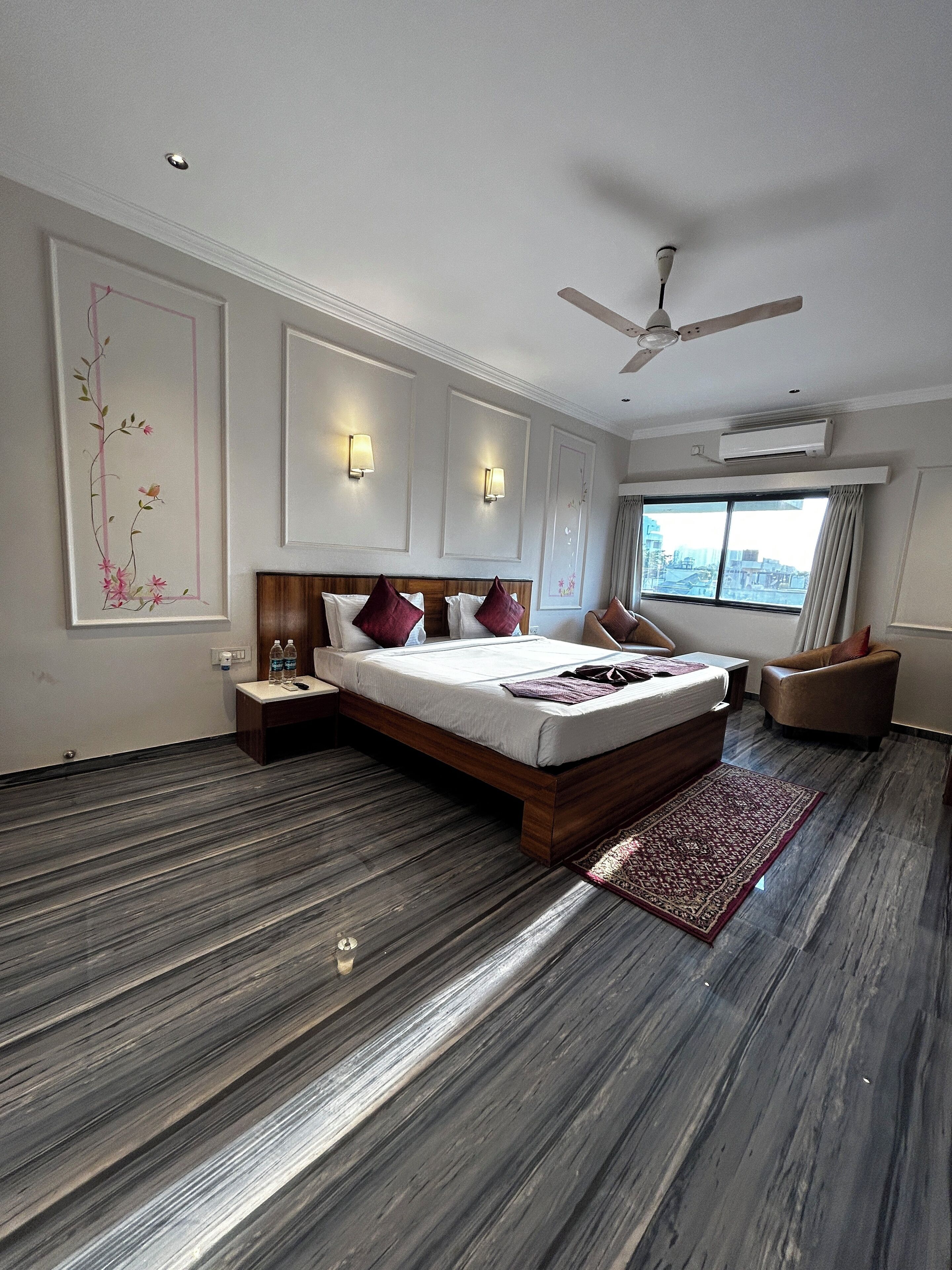 A modern hotel room with a large bed, a ceiling fan, a nightstand, a chair, and a view of the city through the window.