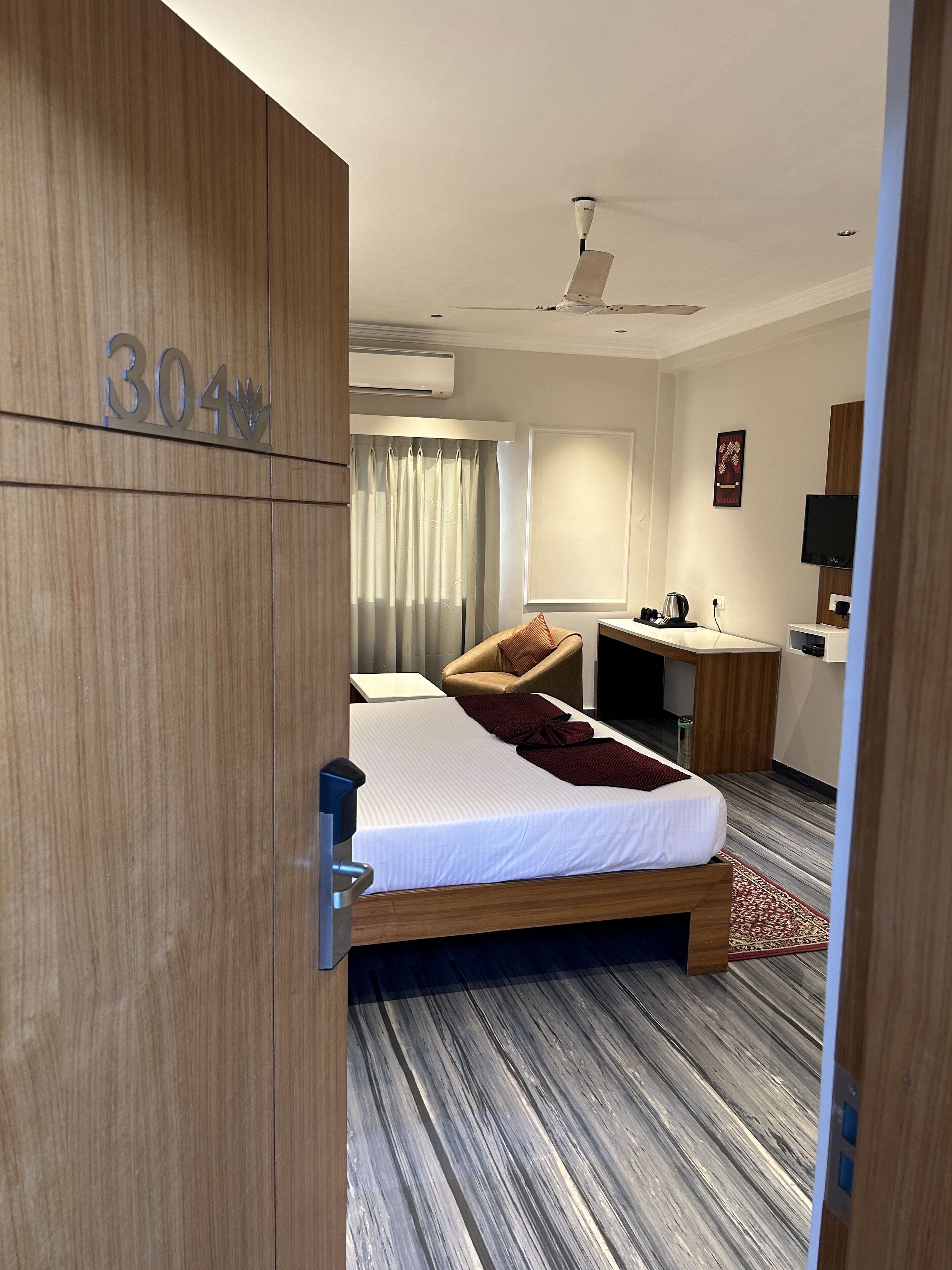 Quarto de hotel com cama, escrivaninha, cadeira, ar condicionado, ventilador de teto e televisão.