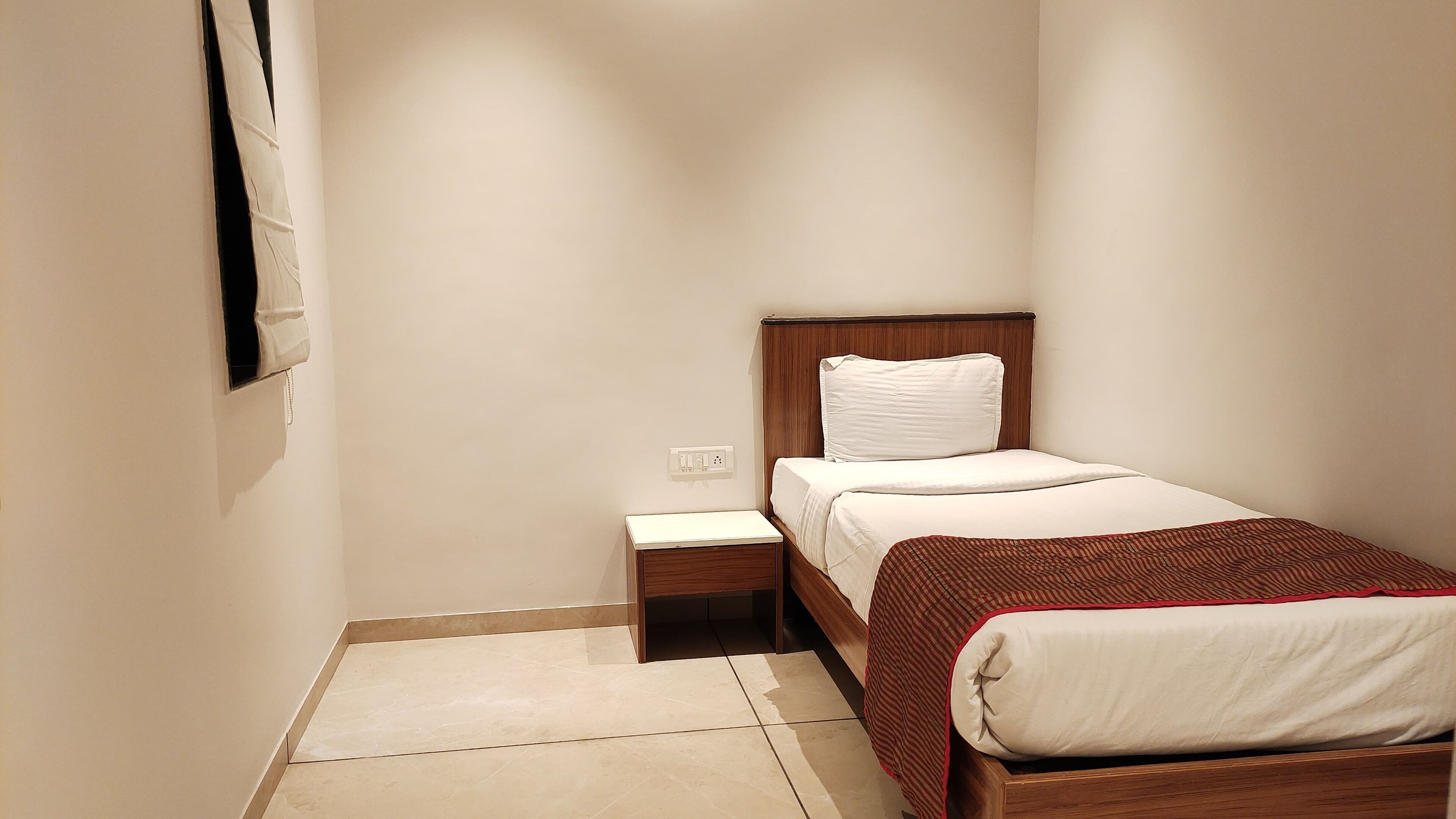 A small, neatly arranged bedroom with a single bed, a bedside table, and a power outlet on the wall.