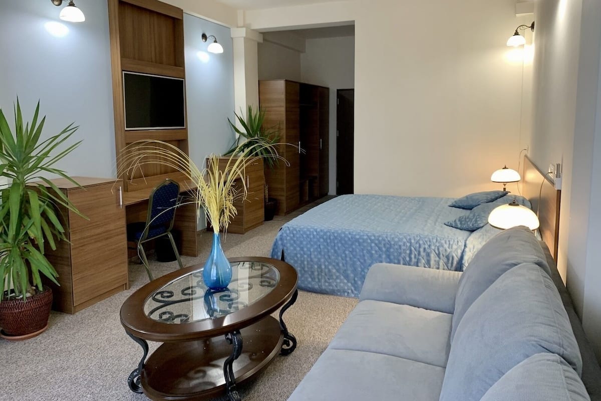 A modern hotel room with a bed, a sofa, a small table with a vase, and two potted plants.