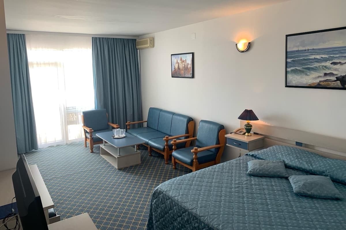 A hotel room with a bed, a sofa, a desk, and a chair. There is a painting on the wall and a lamp on the desk.