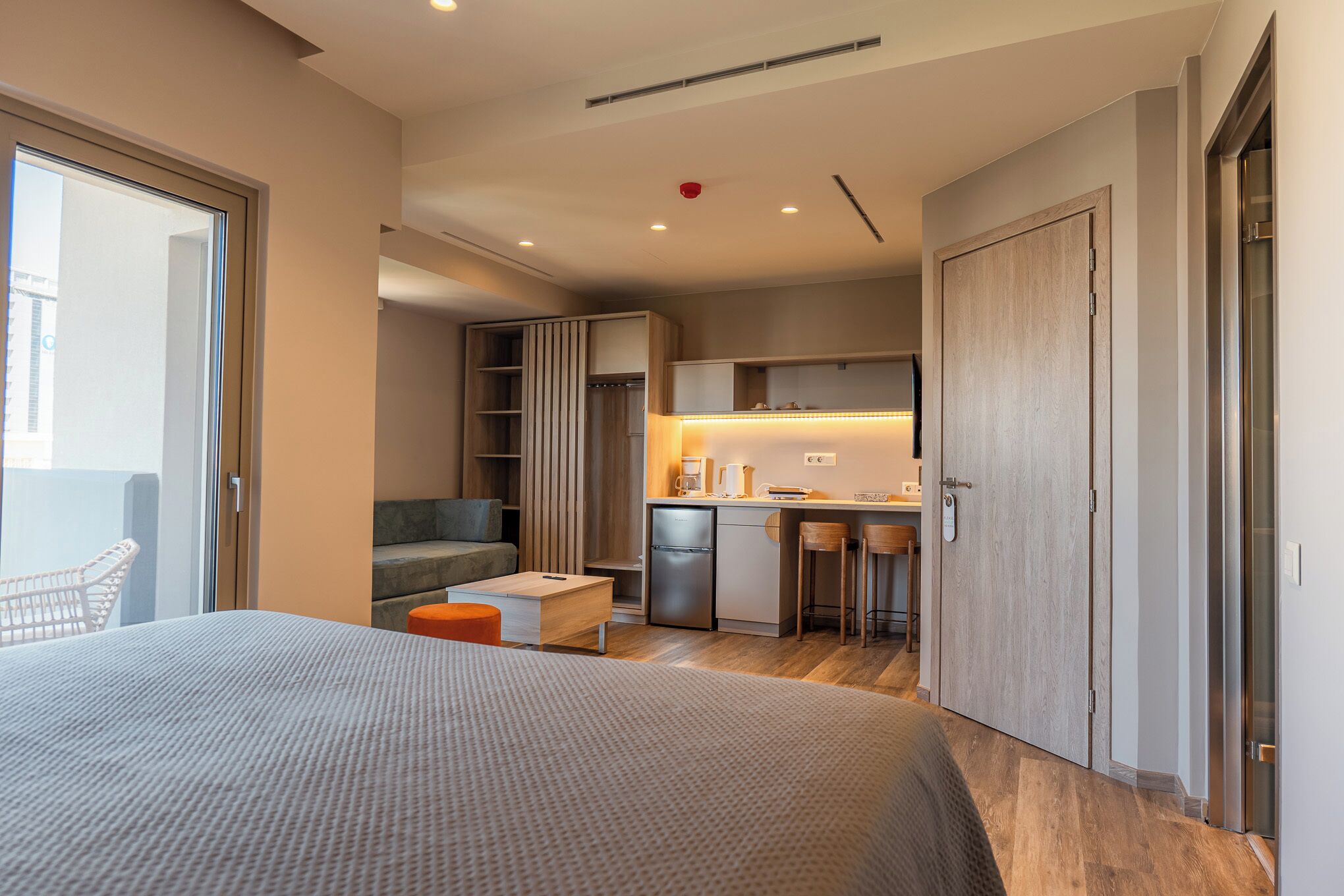 Una habitación de hotel moderna con una cama grande, un escritorio pequeño, una kitchenette y un balcón con puerta corrediza.