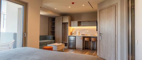Une chambre d’hôtel moderne dotée d’un grand lit, d’un petit bureau, d’une kitchenette et d’une porte coulissante donnant sur un balcon.
