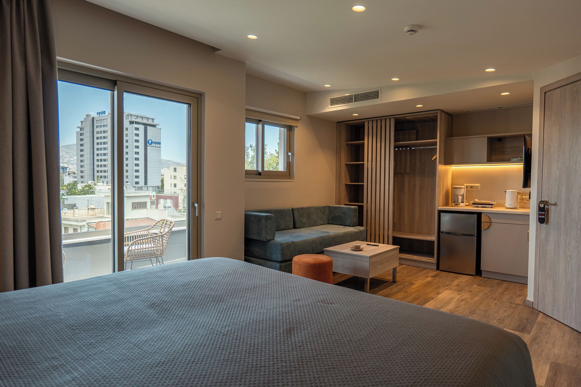 A modern hotel room with a large bed, a seating area with a sofa and armchair, a small table, a kitchenette, and a view of the city through large windows.