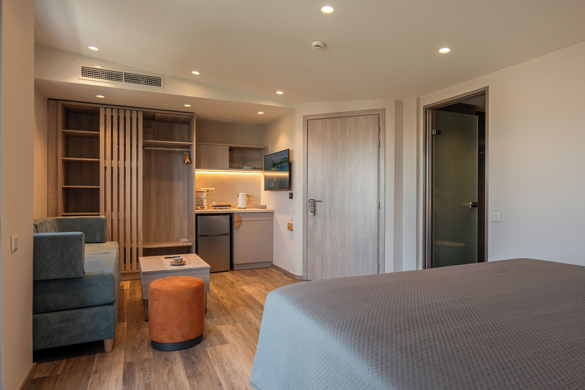 A compact hotel room with a bed, a sofa, a small table, a kitchenette, and a wardrobe.