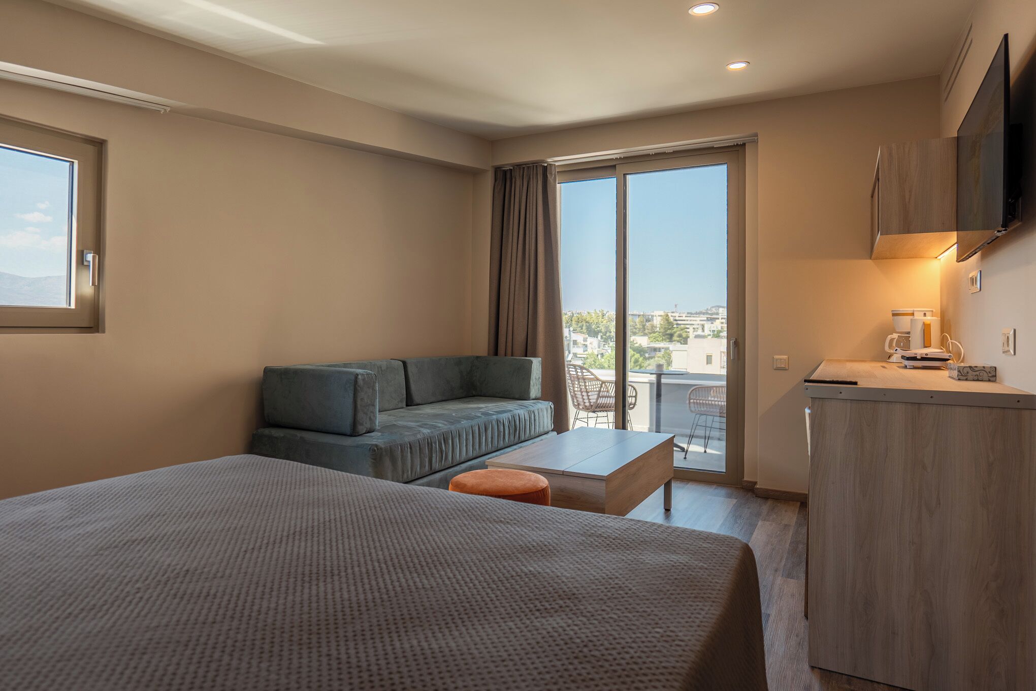 A modern hotel room with a large bed, a sofa, a small table, a coffee machine, and a TV mounted on the wall.