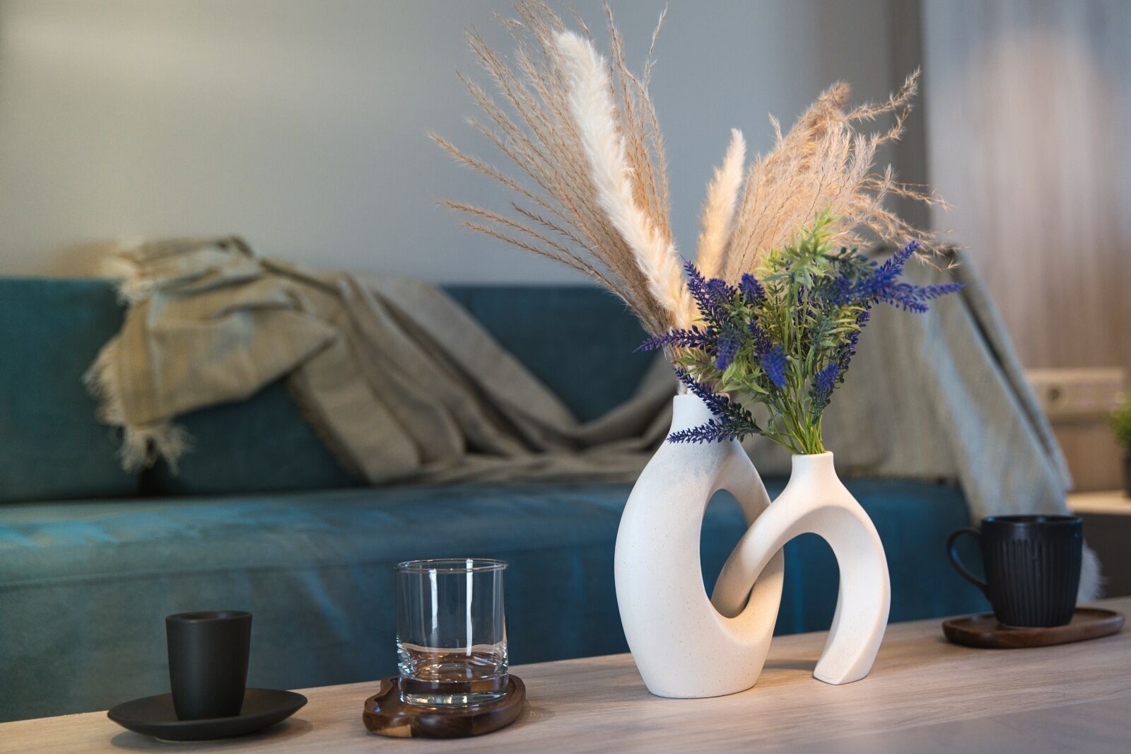 Ein modernes Wohnzimmer mit einem blauen Sofa, zwei weißen Vasen mit Trockenpflanzen, einem Glas Wasser und einer schwarzen Tasse auf einem Holztisch.