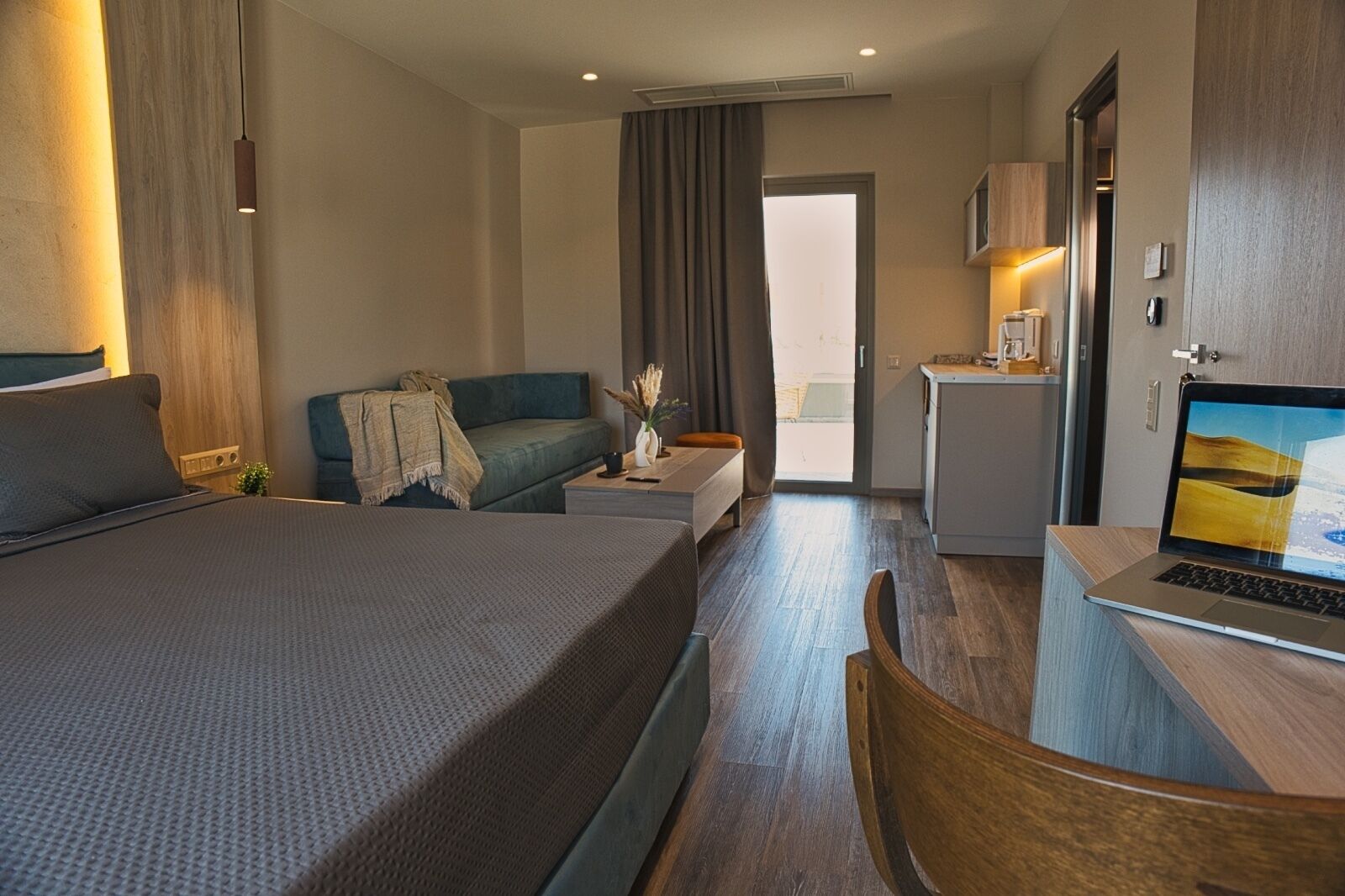 Ein Hotelzimmer mit einem Bett, einer Couch, einem Schreibtisch mit einem Laptop, einer kleinen Küchenzeile und Blick ins Freie.