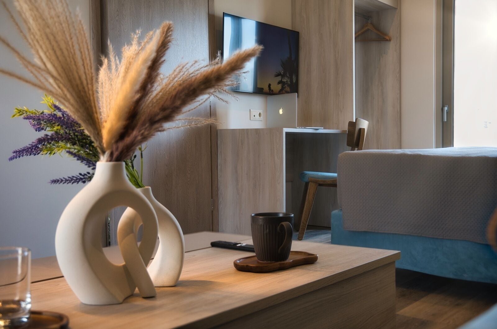 A modern living room with a minimalist design, featuring a white vase with dried pampas grass and purple flowers, a dark mug on a wooden coaster, and a flat-screen TV mounted on the wall.