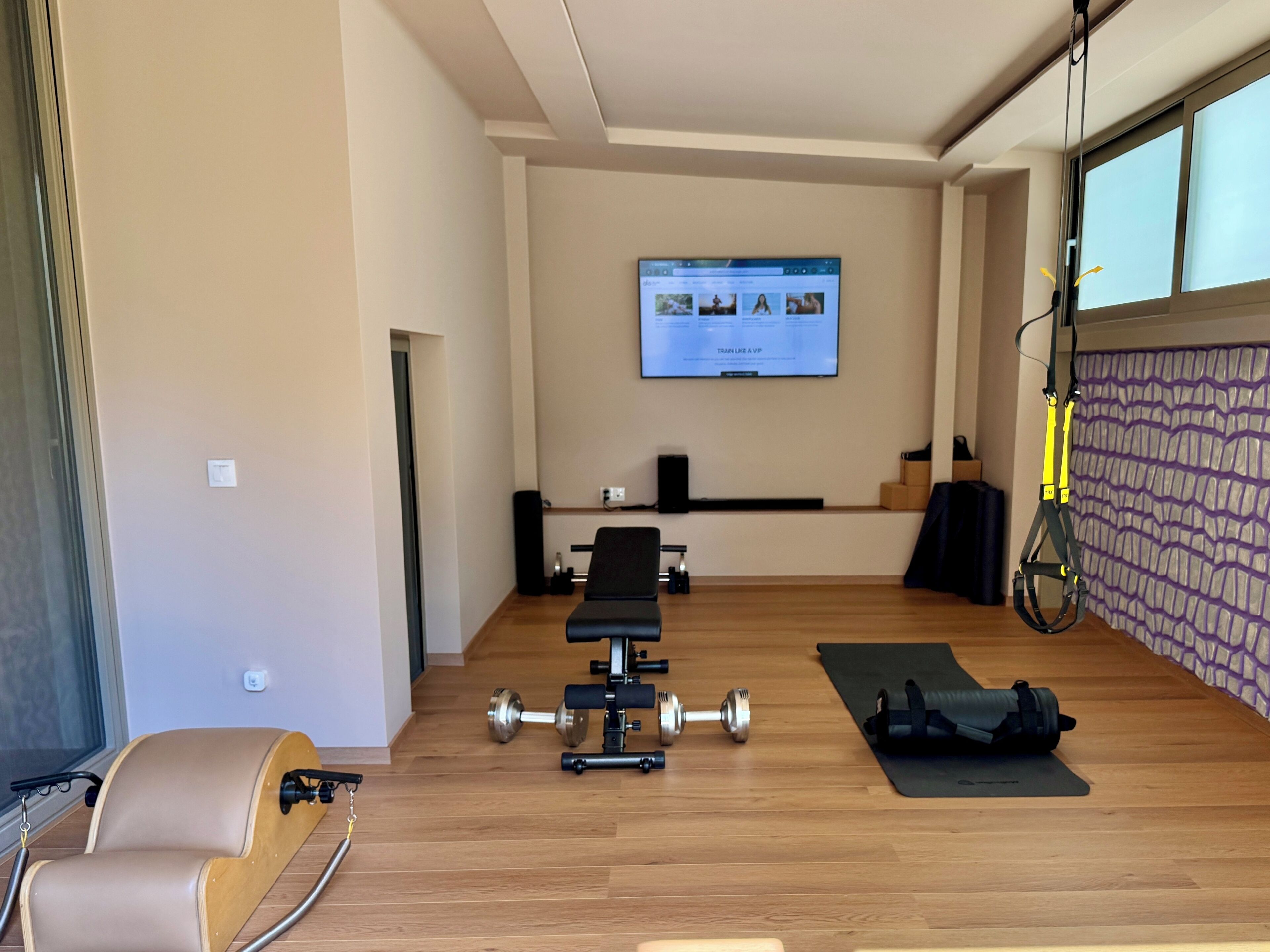 Uma academia moderna em casa, com equipamentos de ginástica, uma TV exibindo um programa de fitness e uma janela grande.