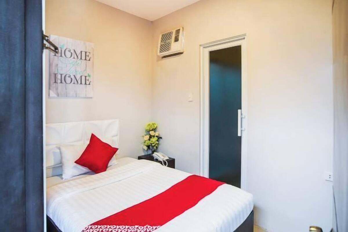 Habitación de hotel con cama, mesita de noche con teléfono, rejilla de ventilación en la pared y una puerta.