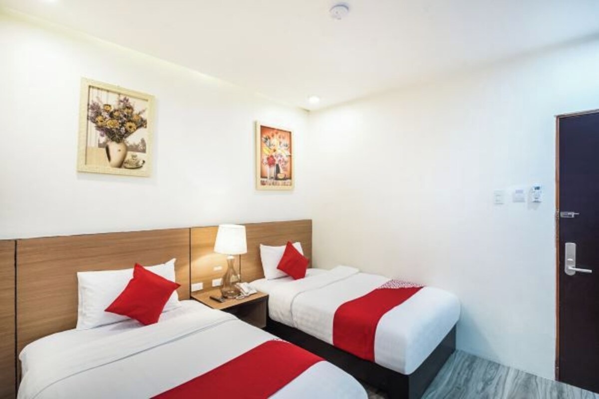 Habitación de hotel con dos camas, cabecero de madera, mesita de noche con lámpara y dos cuadros colgados en la pared.