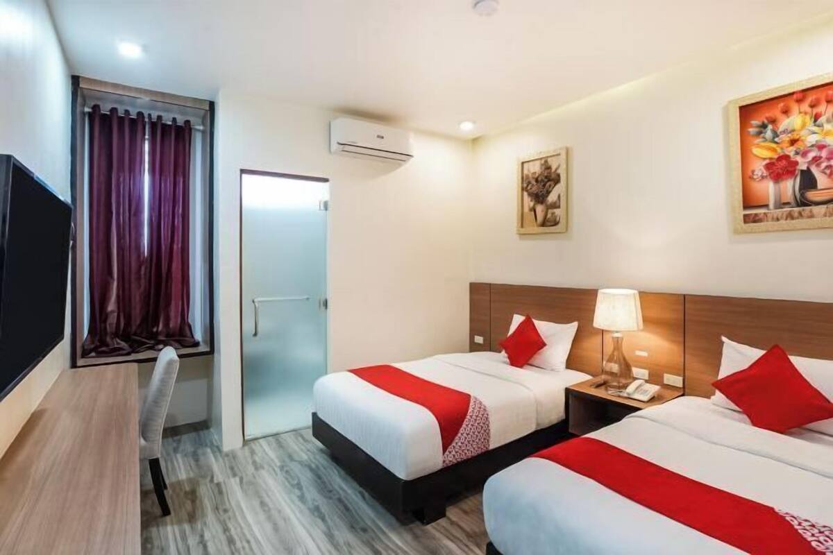 Habitación de hotel con dos camas, un escritorio, una silla, un equipo de aire acondicionado y un cuadro en la pared.