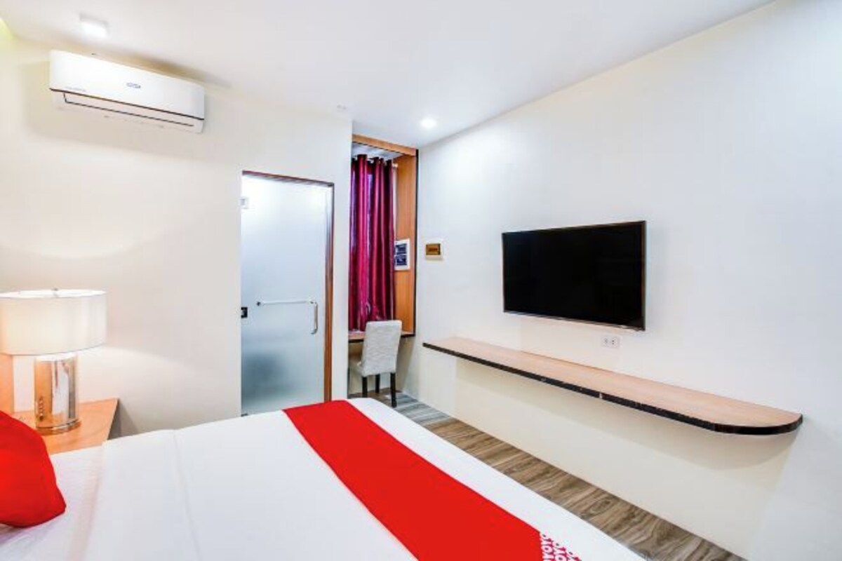 Una habitación de hotel con una cama, un televisor montado en la pared, una mesita de noche con lámpara y un detalle rojo en la colcha.