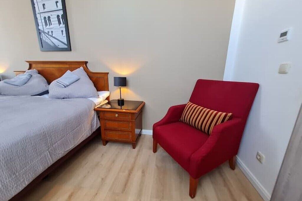 A hotel room with a bed, a nightstand with a lamp, a red armchair, and a framed picture on the wall.
