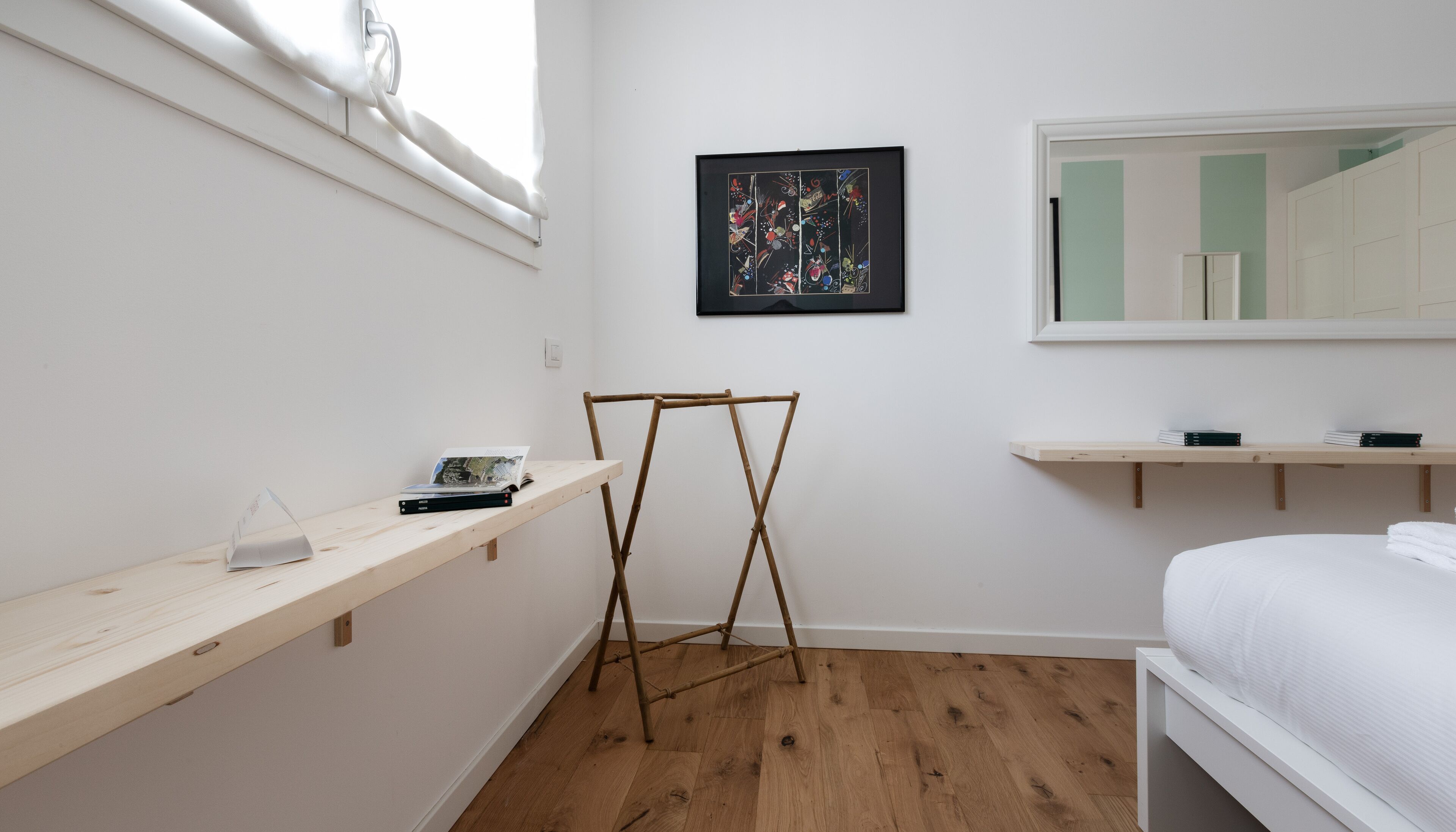 Ein minimalistisches Schlafzimmer mit Holzboden, einem Bett mit weißer Bettwäsche, einem Holzregal mit Büchern, einem gerahmten abstrakten Gemälde und einem Fenster mit weißen Vorhängen.