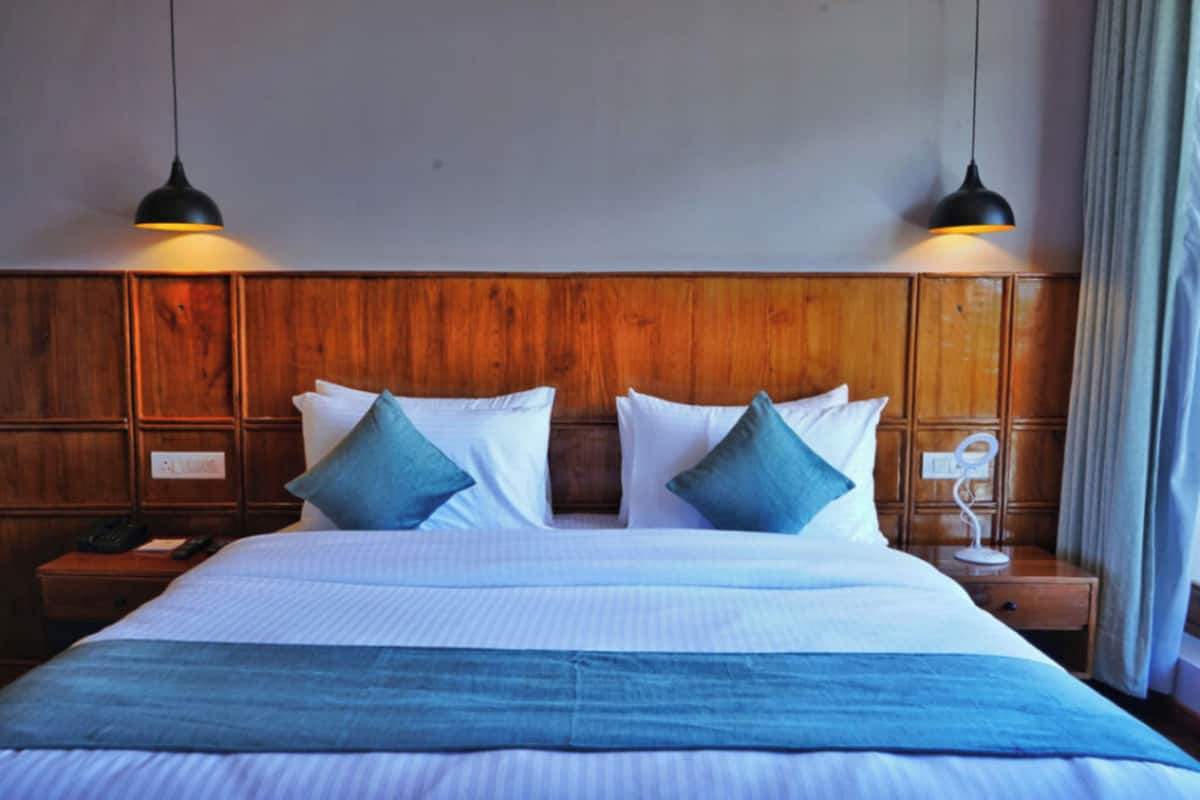 Habitación de hotel con cabecera de madera, una cama grande con ropa de cama blanca y azul, dos lámparas de noche y una mesita de noche con un teléfono y una pequeña mesa.